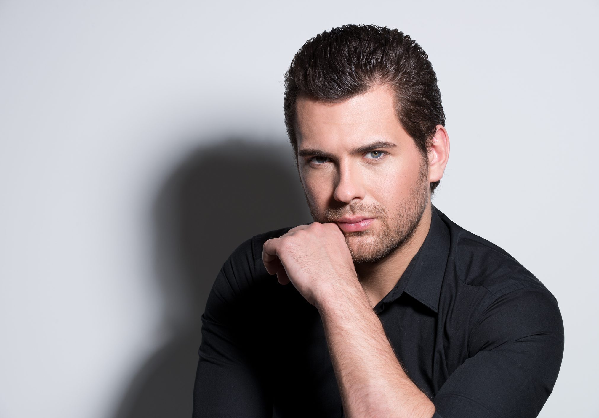 Fashion portrait of handsome man in black shirt with hand near face poses in the studio.